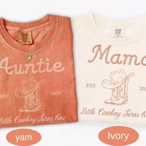 May include: Two crew neck t-shirts in yam and ivory colors. The yam shirt features white text reading Auntie, a cowboy hat and boot graphic, and Little Cowboy Turns One. The ivory shirt features brown text reading Mama, the same graphic, and Little Cowboy Turns One.