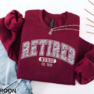 May include: Maroon crewneck sweatshirt with the text "RETIRED NURSE EST. 2026" in a floral design. A silver necklace with a pendant is draped on the sweatshirt. The sweatshirt is paired with a pair of blue jeans.