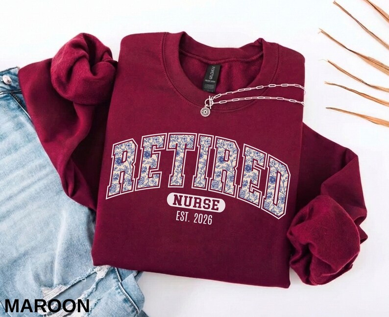 May include: Maroon crewneck sweatshirt with the text "RETIRED NURSE EST. 2026" in a floral design. A silver necklace with a pendant is draped on the sweatshirt. The sweatshirt is paired with a pair of blue jeans.