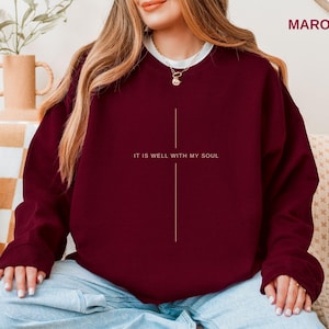 May include: Maroon sweatshirt with the text "IT IS WELL WITH MY SOUL" in white. The sweatshirt has a simple, vertical line design. The model is wearing a white undershirt and light blue jeans. The word "MAROON" is in the top right corner.