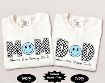 May include: Two ivory t-shirts featuring black and white checkered letters spelling MOM and DAD. The letter O is replaced by a light blue smiley face with black lightning bolt eyes. Below the text, the phrase Oliver's One Happy Dude is printed.