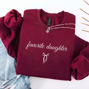 Puede incluir: Una sudadera granate con las palabras "favorite daughter" en escritura blanca y un lazo blanco. Un collar plateado con un colgante est&aacute; sobre la sudadera. La sudadera se combina con unos vaqueros azules.