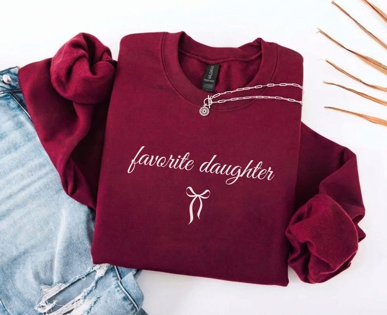 Puede incluir: Una sudadera granate con las palabras "favorite daughter" en escritura blanca y un lazo blanco. Un collar plateado con un colgante est&aacute; sobre la sudadera. La sudadera se combina con unos vaqueros azules.