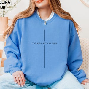 May include: Carolina blue sweatshirt with the text "IT IS WELL WITH MY SOUL" in black. The sweatshirt has a simple cross design. The model is wearing a white undershirt and a necklace.