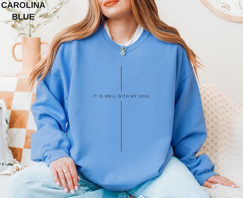 May include: Carolina blue sweatshirt with the text "IT IS WELL WITH MY SOUL" in black. The sweatshirt has a simple cross design. The model is wearing a white undershirt and a necklace.