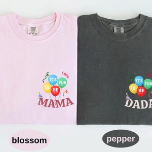 Pu&ograve; includere: Una t-shirt rosa chiaro e una t-shirt grigio scuro, ciascuna con una grafica di palloncini colorati con il testo IT'S FUN TO BE MAMA sulla maglietta rosa e IT'S FUN TO BE DADA su quella grigia, accompagnate da cappellini e festoni.