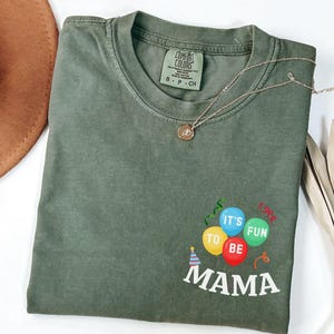 Pu&ograve; includere: Una t-shirt a maniche corte color verde oliva con scollo rotondo. La parte anteriore presenta una grafica con tre palloncini gialli, blu e verdi, con la scritta 'IT'S FUN TO BE MAMA' stampata in bianco sotto, e un piccolo cappellino da festa.