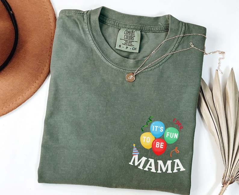 Pu&ograve; includere: Una t-shirt a maniche corte color verde oliva con scollo rotondo. La parte anteriore presenta una grafica con tre palloncini gialli, blu e verdi, con la scritta 'IT'S FUN TO BE MAMA' stampata in bianco sotto, e un piccolo cappellino da festa.