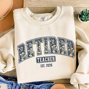 May include: A sand-colored sweatshirt with the word "RETIRED" in navy blue, floral-filled letters. Below, the text "TEACHER EST. 2026" is displayed. The sweatshirt is folded, with a pair of blue jeans and a hat visible.