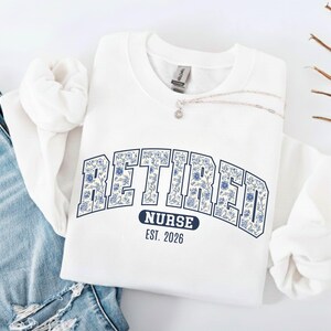 May include: White sweatshirt with the word "RETIRED" in a floral design, arched across the chest. Below the word "RETIRED" is the word "NURSE" and the year "EST. 2026" in a navy blue banner. A silver necklace is draped across the sweatshirt.
