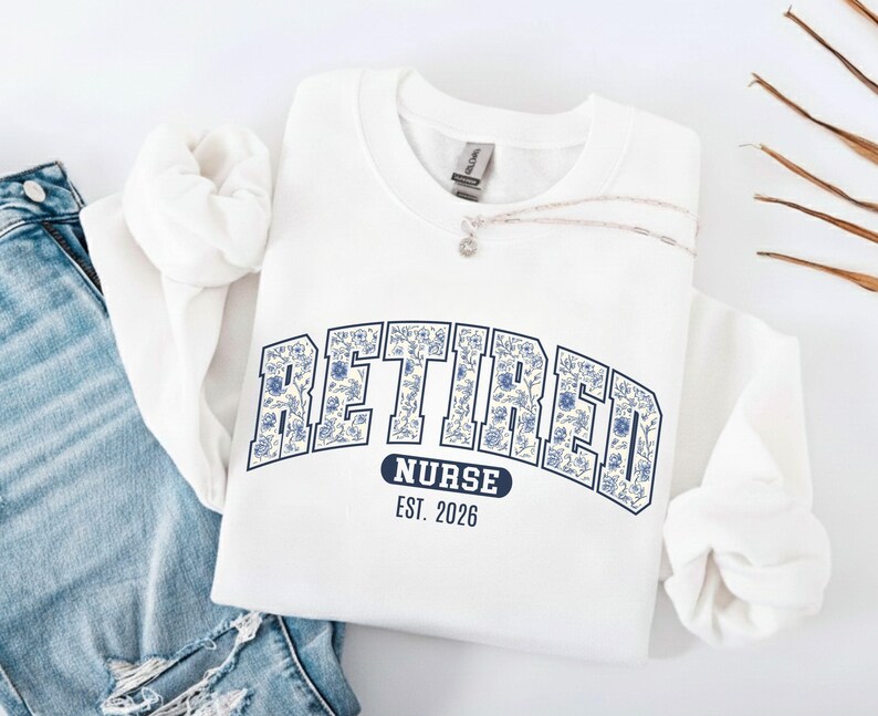 May include: White sweatshirt with the word "RETIRED" in a floral design, arched across the chest. Below the word "RETIRED" is the word "NURSE" and the year "EST. 2026" in a navy blue banner. A silver necklace is draped across the sweatshirt.