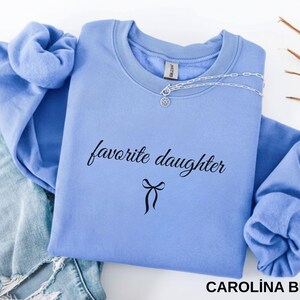 Puede incluir: Sudadera azul Carolina con las palabras "favorite daughter" en escritura negra y un lazo. Un collar plateado est&aacute; colocado en la parte superior. La parte superior de manga larga est&aacute; doblada sobre un fondo claro.