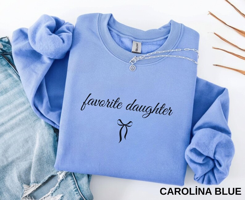 Puede incluir: Sudadera azul Carolina con las palabras "favorite daughter" en escritura negra y un lazo. Un collar plateado est&aacute; colocado en la parte superior. La parte superior de manga larga est&aacute; doblada sobre un fondo claro.