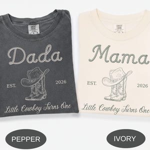May include: Two crew neck t-shirts in pepper and ivory colors. The pepper shirt features white text reading Dada, EST. 2026, and Little Cowboy Turns One with a cowboy hat and boot graphic. The ivory shirt features dark text with the same design.