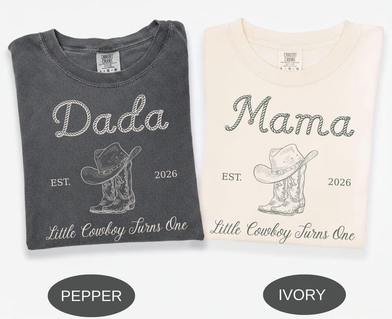 May include: Two crew neck t-shirts in pepper and ivory colors. The pepper shirt features white text reading Dada, EST. 2026, and Little Cowboy Turns One with a cowboy hat and boot graphic. The ivory shirt features dark text with the same design.