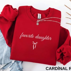 Puede incluir: Una sudadera roja cardenal con las palabras "favorite daughter" en escritura cursiva y un gr&aacute;fico de lazo. Un collar plateado est&aacute; drapeado en la parte superior. La sudadera est&aacute; doblada junto a unos vaqueros azules.