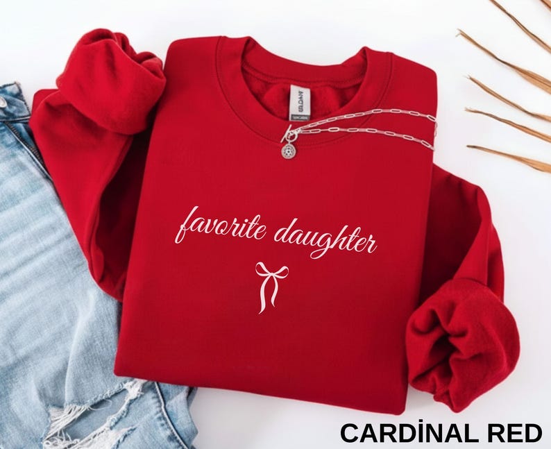 Puede incluir: Una sudadera roja cardenal con las palabras "favorite daughter" en escritura cursiva y un gr&aacute;fico de lazo. Un collar plateado est&aacute; drapeado en la parte superior. La sudadera est&aacute; doblada junto a unos vaqueros azules.