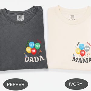 Pu&ograve; includere: Una t-shirt color pepe e una t-shirt color avorio, entrambe con una grafica di palloncini colorati con il testo 'IT'S FUN TO BE DADA' sulla maglia color pepe e 'IT'S FUN TO BE MAMA' su quella avorio, accompagnate da festoni e un cappellino.