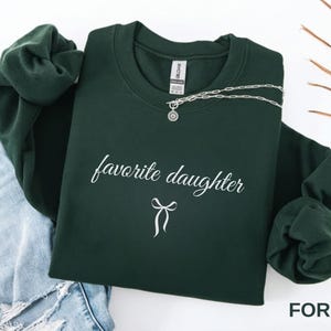 Puede incluir: Una sudadera verde oscuro con la frase "favorite daughter" en escritura cursiva y un lazo. Un collar plateado con un colgante est&aacute; colocado sobre la sudadera. La palabra "FOREST" es visible en la esquina inferior derecha.