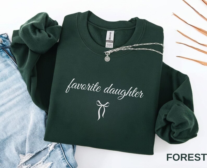 Puede incluir: Una sudadera verde oscuro con la frase "favorite daughter" en escritura cursiva y un lazo. Un collar plateado con un colgante est&aacute; colocado sobre la sudadera. La palabra "FOREST" es visible en la esquina inferior derecha.