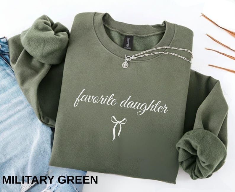 Puede incluir: Sudadera verde militar con el texto "favorite daughter" en escritura blanca. Un collar plateado con un colgante est&aacute; drapeado en la parte delantera. La sudadera est&aacute; doblada, con las mangas a la vista.