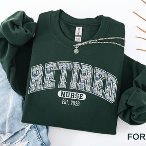 May include: A forest green sweatshirt with the word "RETIRED" in a floral design, "NURSE" and "EST. 2026" printed below. A silver necklace with a pendant rests on the sweatshirt. The sweatshirt is folded next to a pair of blue jeans.