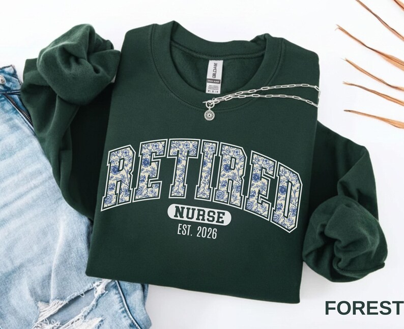 May include: A forest green sweatshirt with the word "RETIRED" in a floral design, "NURSE" and "EST. 2026" printed below. A silver necklace with a pendant rests on the sweatshirt. The sweatshirt is folded next to a pair of blue jeans.