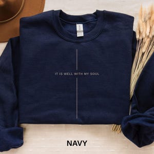 May include: Navy blue crewneck sweatshirt with the text "IT IS WELL WITH MY SOUL" in white, centered vertically. A white vertical line bisects the text. The word "NAVY" is printed in white at the bottom. The sweatshirt is folded.
