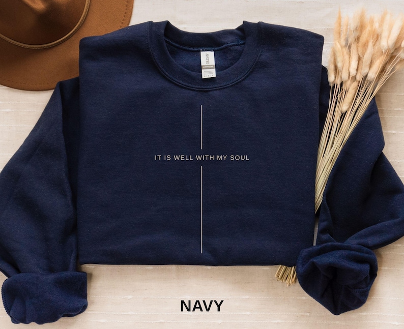 May include: Navy blue crewneck sweatshirt with the text "IT IS WELL WITH MY SOUL" in white, centered vertically. A white vertical line bisects the text. The word "NAVY" is printed in white at the bottom. The sweatshirt is folded.