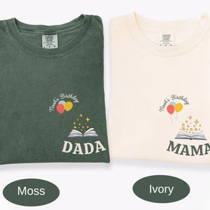 May include: Two t-shirts, one moss green and one ivory, are displayed. Each shirt has a small graphic of balloons, a book, and text. The moss green shirt says "DADA", and the ivory shirt says "MAMA".