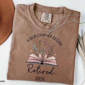 May include: Espresso-colored t-shirt with the text "A new chapter begins" and "Retired 2026". The shirt features an open book graphic with floral accents. A silver necklace is draped across the shirt.