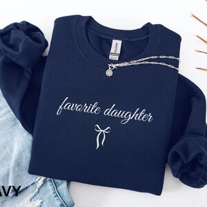 Puede incluir: Sudadera azul marino con la frase "favorite daughter" en escritura cursiva y un lazo gr&aacute;fico. Un collar plateado con un colgante est&aacute; colocado en la parte superior. La palabra "NAVY" est&aacute; impresa en la parte inferior izquierda.
