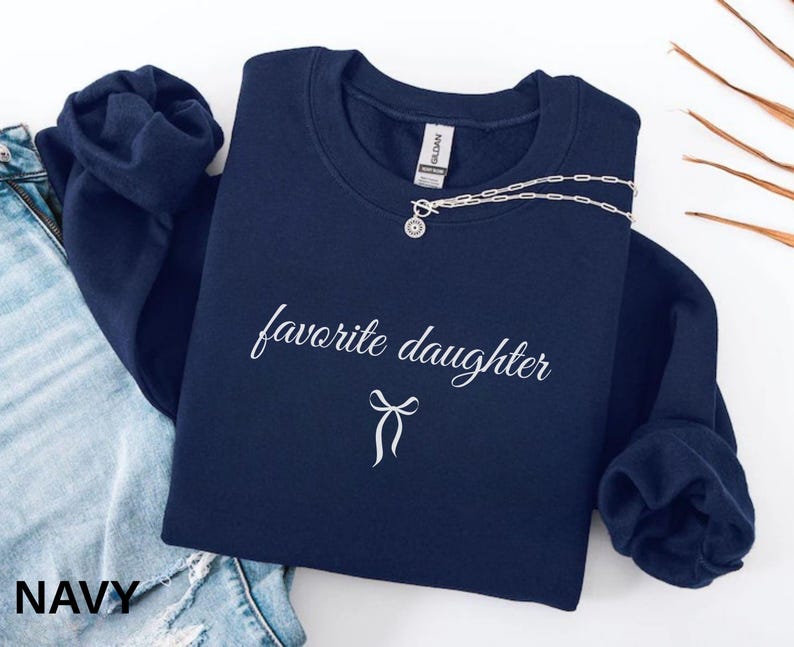 Puede incluir: Sudadera azul marino con la frase "favorite daughter" en escritura cursiva y un lazo gr&aacute;fico. Un collar plateado con un colgante est&aacute; colocado en la parte superior. La palabra "NAVY" est&aacute; impresa en la parte inferior izquierda.