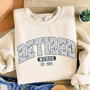 May include: A sand-colored sweatshirt with the word "RETIRED" in large, blue floral letters. Below, it reads "NURSE EST. 2026" in a smaller, navy blue font. The sweatshirt has a crew neck and long sleeves.