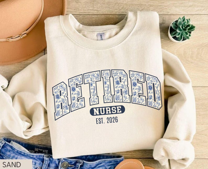 May include: A sand-colored sweatshirt with the word "RETIRED" in large, blue floral letters. Below, it reads "NURSE EST. 2026" in a smaller, navy blue font. The sweatshirt has a crew neck and long sleeves.