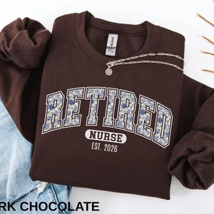May include: A dark chocolate-colored sweatshirt with the text "RETIRED NURSE EST. 2026" in a collegiate-style font with a floral pattern. A silver necklace with a pendant is draped across the sweatshirt. The sleeves are folded.