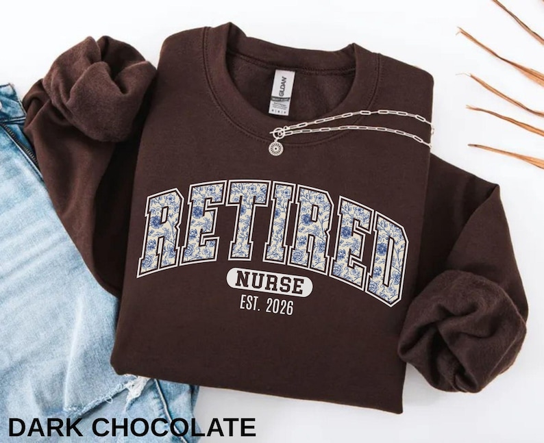 May include: A dark chocolate-colored sweatshirt with the text "RETIRED NURSE EST. 2026" in a collegiate-style font with a floral pattern. A silver necklace with a pendant is draped across the sweatshirt. The sleeves are folded.
