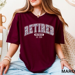 May include: Maroon t-shirt with the word "RETIRED" in a floral design, arched across the chest. Below the word "RETIRED" is the word "NURSE" and the year "EST. 2026". The shirt is a short-sleeved crew neck.