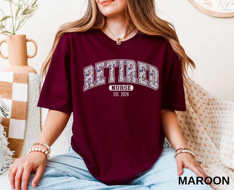 May include: Maroon t-shirt with the word "RETIRED" in a floral design, arched across the chest. Below the word "RETIRED" is the word "NURSE" and the year "EST. 2026". The shirt is a short-sleeved crew neck.