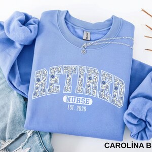 May include: Carolina blue sweatshirt with the word "RETIRED" in white, floral-filled letters. Below, it reads "NURSE EST. 2026". A silver necklace with a pendant is draped across the neckline. The sweatshirt is folded on top of a pair of blue jeans.