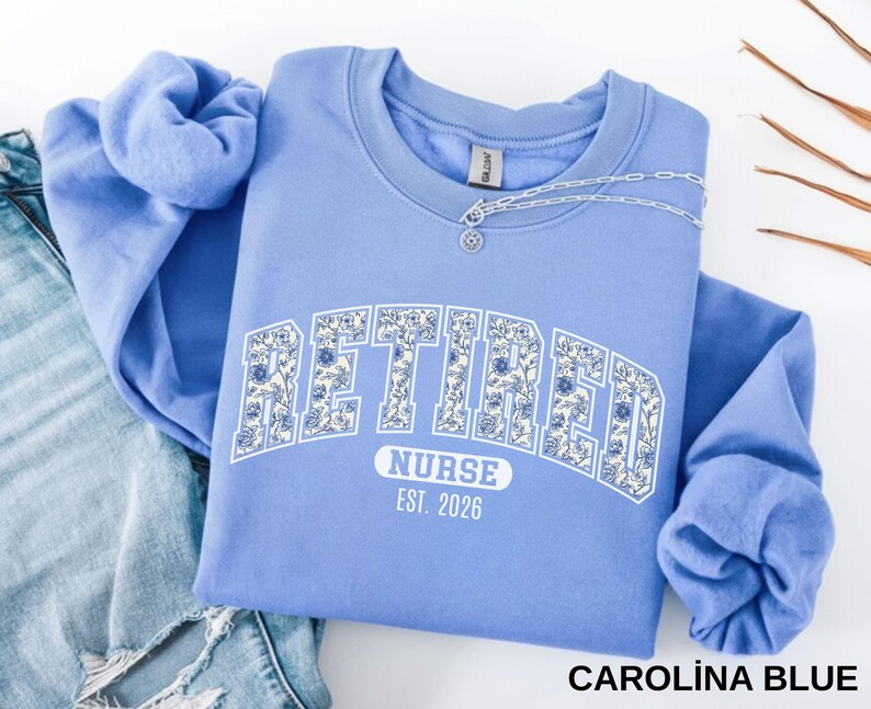 May include: Carolina blue sweatshirt with the word "RETIRED" in white, floral-filled letters. Below, it reads "NURSE EST. 2026". A silver necklace with a pendant is draped across the neckline. The sweatshirt is folded on top of a pair of blue jeans.