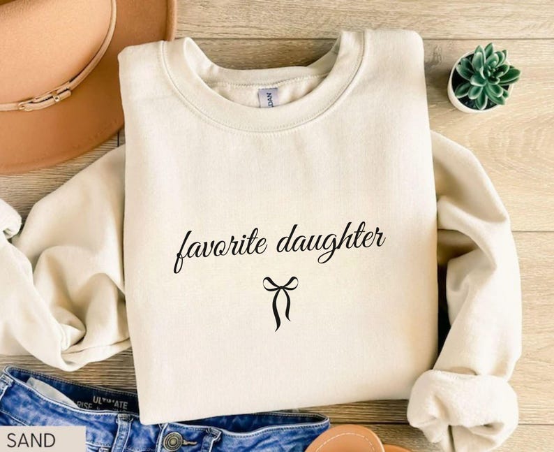 Puede incluir: Una sudadera color arena con la frase "favorite daughter" en fuente negra y un lazo negro. La sudadera est&aacute; doblada sobre una superficie de madera, con unos vaqueros azules y un sombrero marr&oacute;n en el fondo.