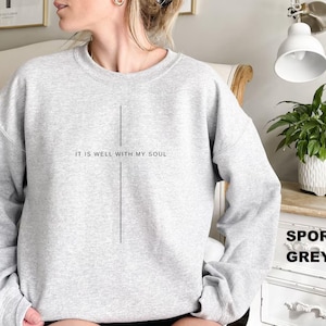 May include: A light gray sweatshirt with the text "IT IS WELL WITH MY SOUL" printed in black. A vertical line design is centered below the text. The sweatshirt is labeled "SPORT GREY".
