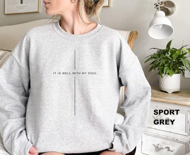 May include: A light gray sweatshirt with the text "IT IS WELL WITH MY SOUL" printed in black. A vertical line design is centered below the text. The sweatshirt is labeled "SPORT GREY".