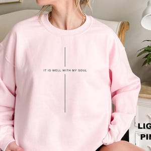 May include: Light pink sweatshirt with the text "IT IS WELL WITH MY SOUL" in black. The sweatshirt has a simple, minimalist design, with a vertical line below the text. The sleeves are long and the material appears soft.
