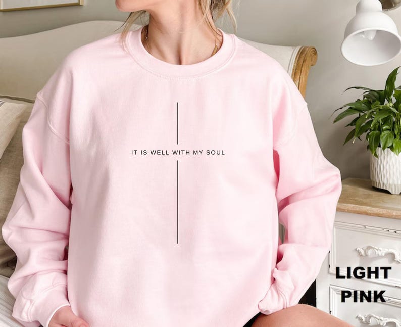 May include: Light pink sweatshirt with the text "IT IS WELL WITH MY SOUL" in black. The sweatshirt has a simple, minimalist design, with a vertical line below the text. The sleeves are long and the material appears soft.