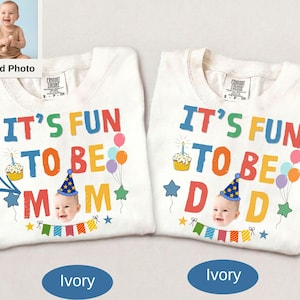 May include: Two ivory t-shirts featuring colourful graphics. One shirt reads IT'S FUN TO BE MOM, the other IT'S FUN TO BE DAD. Both include a cupcake, a baby's face wearing a blue party hat, balloons, stars, and a colourful pennant banner.