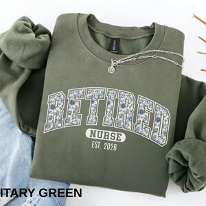 May include: Military green sweatshirt with the word "RETIRED" in a floral design, arched over the word "NURSE" and the year "EST. 2026". A silver necklace is draped over the sweatshirt. The sweatshirt is folded over a pair of blue jeans.