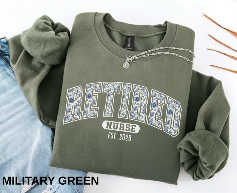 May include: Military green sweatshirt with the word "RETIRED" in a floral design, arched over the word "NURSE" and the year "EST. 2026". A silver necklace is draped over the sweatshirt. The sweatshirt is folded over a pair of blue jeans.