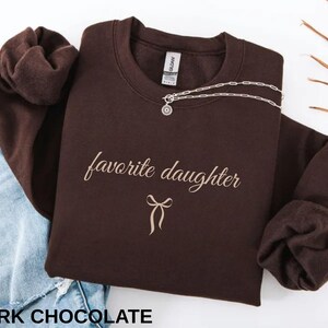 Puede incluir: Una sudadera de color chocolate oscuro con las palabras "favorite daughter" en una fuente cursiva, junto con un gr&aacute;fico de lazo. Un collar plateado con un colgante est&aacute; drapeado en la parte delantera. El texto "DARK CHOCOLATE" est&aacute; en la parte inferior.